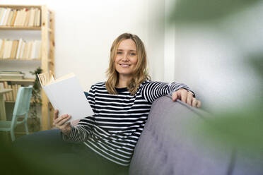 Blond woman smiling while sitting with book on sofa - GIOF11993