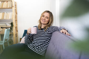 Smiling woman with coffee cup on sofa at home - GIOF11994