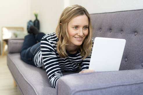 Lächelnde Frau, die ein digitales Tablet benutzt, während sie zu Hause auf dem Sofa liegt - GIOF11995