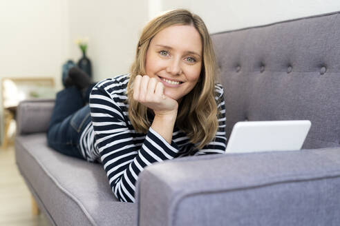 Smiling blond woman with digital tablet lying on sofa at home - GIOF11996