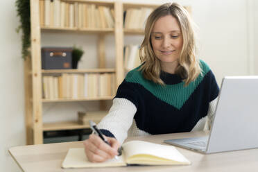Lächelnde Frau schreibt in ein Buch, während sie zu Hause am Laptop sitzt - GIOF11999
