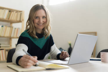 Blond woman smiling while sitting by laptop on table at home - GIOF12000