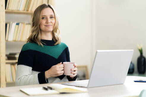 Blonde Frau mit Kaffeetasse und Laptop zu Hause - GIOF12002