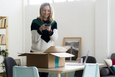 Lächelnde Frau, die ein Foto von einem Paketkarton durch ein Mobiltelefon zu Hause macht - GIOF12014