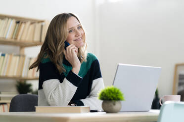 Smiling woman talking on mobile phone while sitting by laptop at home - GIOF12016