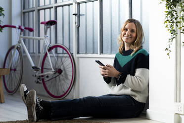 Mittlere erwachsene Frau mit Mobiltelefon, die zu Hause am Fenster sitzt - GIOF12021