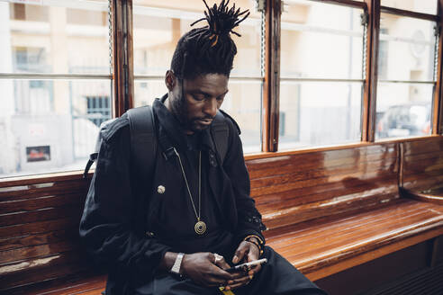 Young man with locs hairstyle using mobile phone sitting in tram - MEUF02329