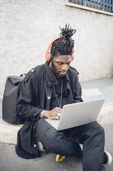 Männlicher Freiberufler mit Kopfhörern, der auf dem Gehweg sitzend einen Laptop benutzt - MEUF02335