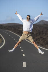 Excited male tourist with arms outstretched jumping over road - SNF01194