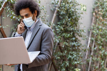 Afro businessman wearing protective face mask holding laptop while talking on smart - IFRF00490