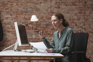 Female professional talking through wireless headphones while holding document at desk - DHEF00589