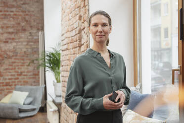 Contented businesswoman holding smart phone while standing by window in office - DHEF00599