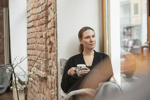 Nachdenkliche Frau mit Teetasse, die zu Hause am Fenster sitzt - DHEF00603