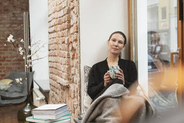 Mature female freelance worker holding tea mug while sitting by window at home - DHEF00604