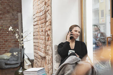 Lächelnde Geschäftsfrau mit Teetasse, die wegschaut, während sie am Fenster mit dem Handy telefoniert - DHEF00605