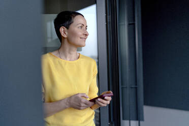 Smiling female entrepreneur day dreaming while holding smart phone at office - OGF01002