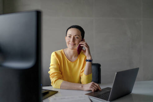 Zufriedene Unternehmerin mit Laptop, die am Schreibtisch in einem modernen Büro mit ihrem Mobiltelefon spricht - OGF01004