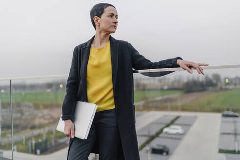 Weibliche Unternehmerin mit Laptop schaut weg, während sie sich auf ein Geländer gegen den Himmel lehnt - OGF01011