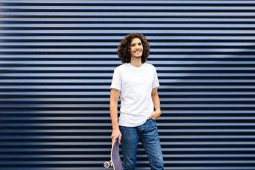 Junger Mann mit lockigem Haar, der ein Skateboard hält, während er vor einem blauen Fensterladen steht - AODF00361