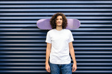 Curly haired man with skateboard behind head while standing in front of shutter - AODF00363