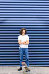 Caucasian young man with arms crossed standing on skateboard in front of blue shutter - AODF00365