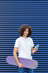 Kaukasischer junger Mann, der ein Smartphone benutzt und ein Skateboard hält - AODF00369