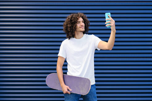 Smiling young man with skateboard taking selfie through smart phone - AODF00371