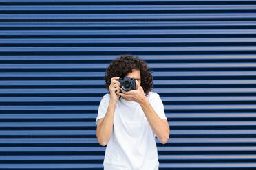 Young man photographing with camera in front of blue shutter - AODF00375