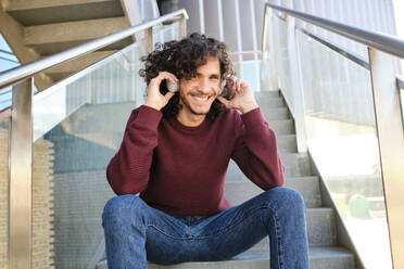 Lächelnder junger Mann mit Kopfhörern, der auf den Stufen eines Bürogebäudes sitzt - AODF00381