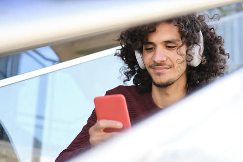 Curly haired young man using smart phone while listening music through headphones - AODF00383