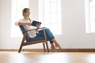 Nachdenkliche Frau schaut weg, während sie ein digitales Tablet auf einem Stuhl am Fenster hält - SBOF03388