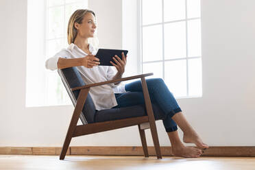 Contemplating woman holding digital tablet while sitting on armchair at home - SBOF03392