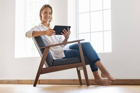 Lächelnde Frau mit digitalem Tablet auf einem Stuhl zu Hause - SBOF03393