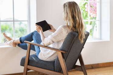 Woman with blond hair holding digital tablet while sitting on armchair at home - SBOF03394