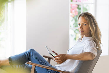 Beautiful woman holding digital tablet while sitting on chair at home - SBOF03398