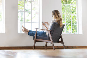 Relaxed woman using smart phone on chair at home - SBOF03401