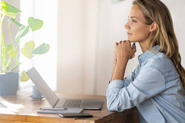 Confident female entrepreneur with hand on chin looking away at home office - SBOF03408