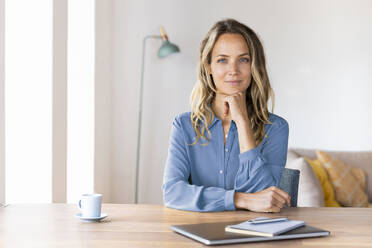 Beautiful businesswoman with hand on chin sitting at desk in home office - SBOF03413