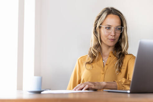 Geschäftsfrau mit Brille, die im Heimbüro auf einen Laptop schaut - SBOF03416