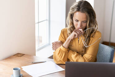 Female professional holding eyeglasses while looking at laptop on desk - SBOF03418