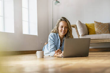 Smiling woman lying on hardwood floor in front of laptop at home - SBOF03420