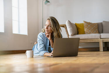 Contemplating woman looking away while lying down with laptop at home - SBOF03421