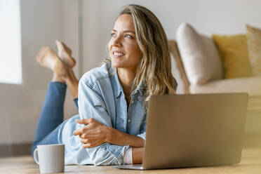 Smiling beautiful woman day dreaming while lying in front of laptop at home - SBOF03422