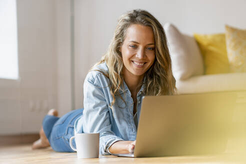 Smiling woman lying on hardwood floor while using laptop at home - SBOF03423