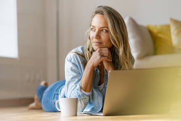 Thoughtful woman with hand on chin looking away while lying on floor in front of laptop - SBOF03424