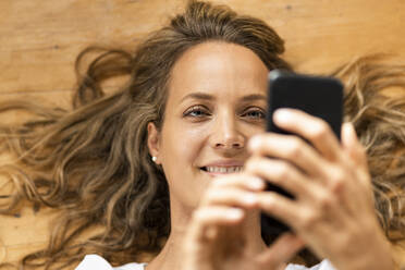 Smiling woman using smart phone while lying on hardwood floor at home - SBOF03429