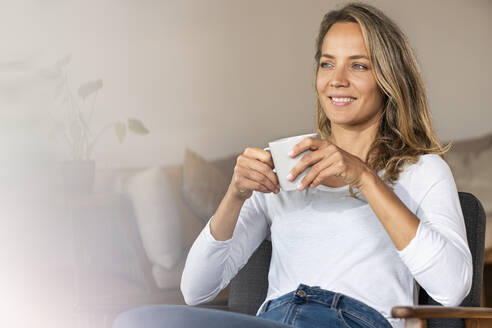 Smiling woman looking away while having coffee at home - SBOF03431