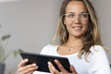 Lächelnde Frau mit Brille, die wegschaut, während sie ein digitales Tablet zu Hause hält - SBOF03433