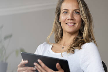 Smiling woman with blue eyes looking away while holding digital tablet at home - SBOF03434