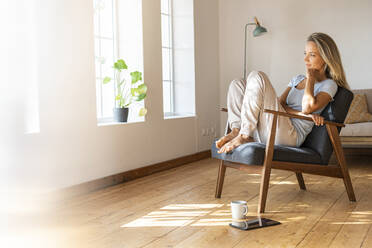 Smiling woman with hand on chin looking away while sitting on chair at home - SBOF03441